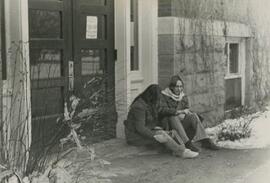 Two people sitting on step outside building.