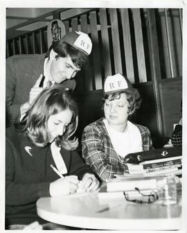 Students at table in beanies writing, 1965