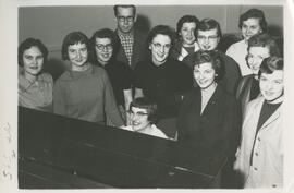 Group of people standing around a piano, no date.