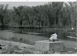 Two people seated on the bank of a river.