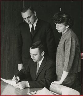 Man signing a paper as others look on, no date.